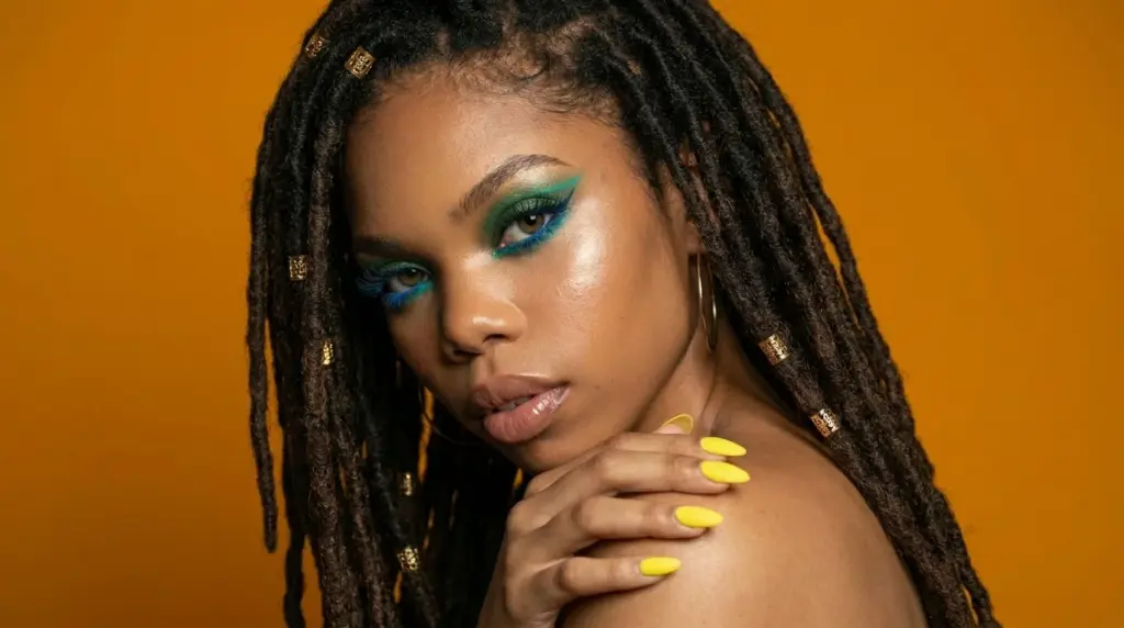 Woman with bold teal and green cruelty-free eyeliner look, featuring dramatic blue lashes and yellow nails against an orange background