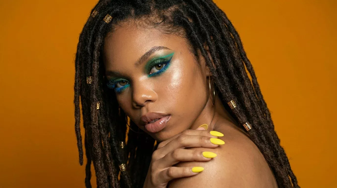Woman with bold teal and green cruelty-free eyeliner look, featuring dramatic blue lashes and yellow nails against an orange background