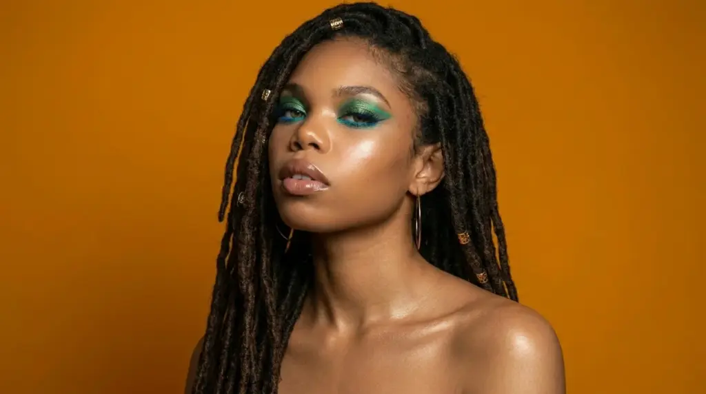Woman with hooded eyes wearing bold teal and green graphic eyeliner look with glowing skin against an orange background