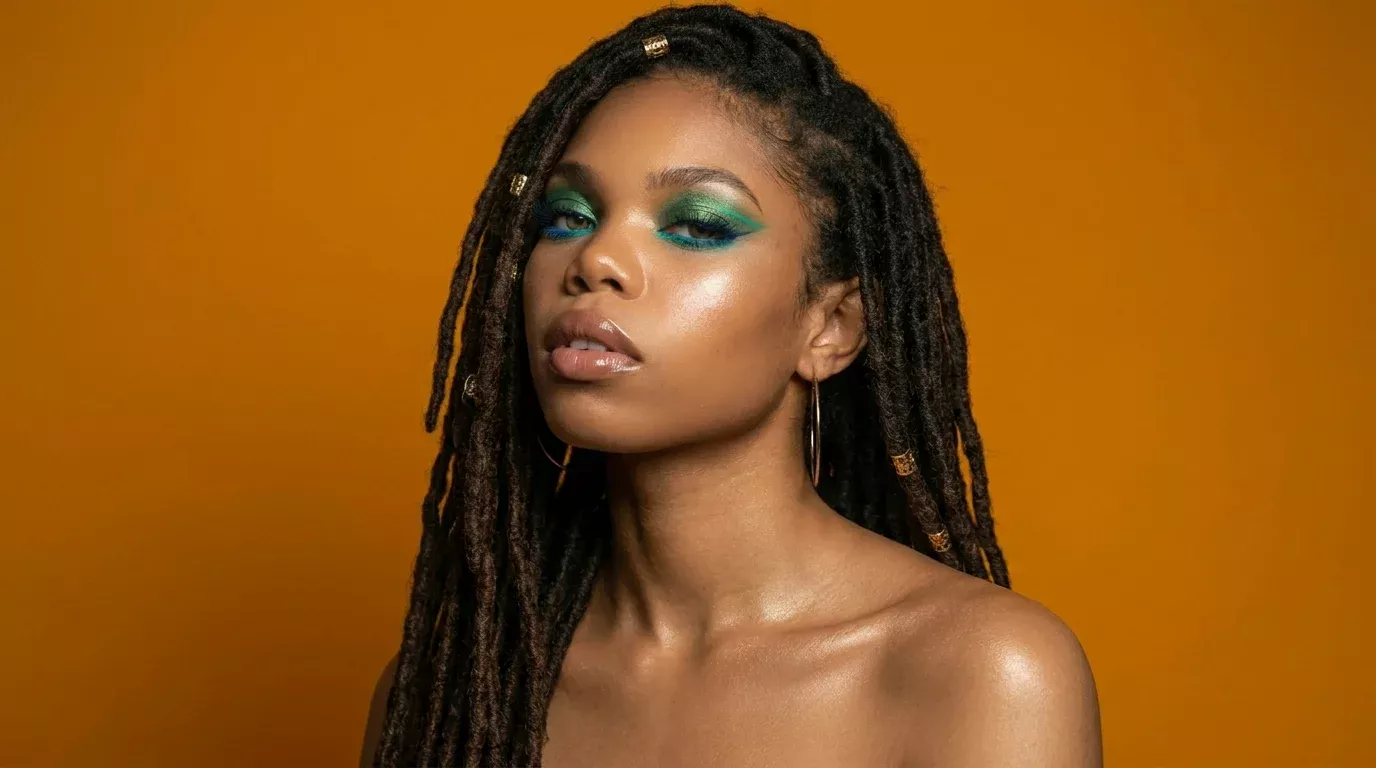 Woman with hooded eyes wearing bold teal and green graphic eyeliner look with glowing skin against an orange background