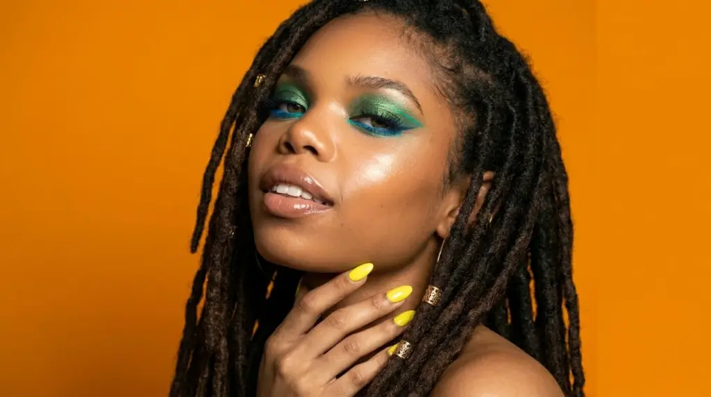 Woman wearing a beginner-friendly teal and green pencil eyeliner look with blue lower liner, yellow nails, and gold rings against an orange background