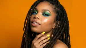 Woman wearing a beginner-friendly teal and green pencil eyeliner look with blue lower liner, yellow nails, and gold rings against an orange background
