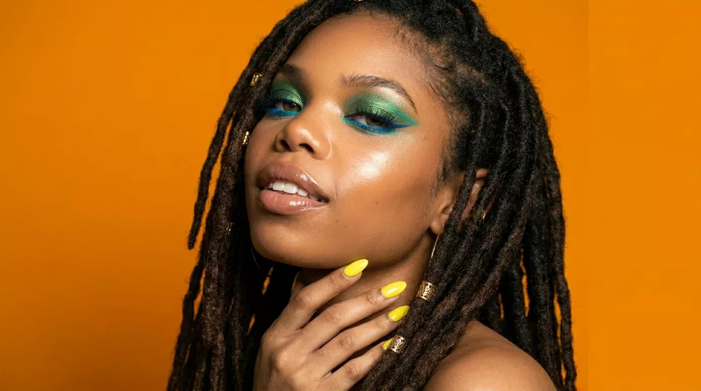 Woman wearing a beginner-friendly teal and green pencil eyeliner look with blue lower liner, yellow nails, and gold rings against an orange background