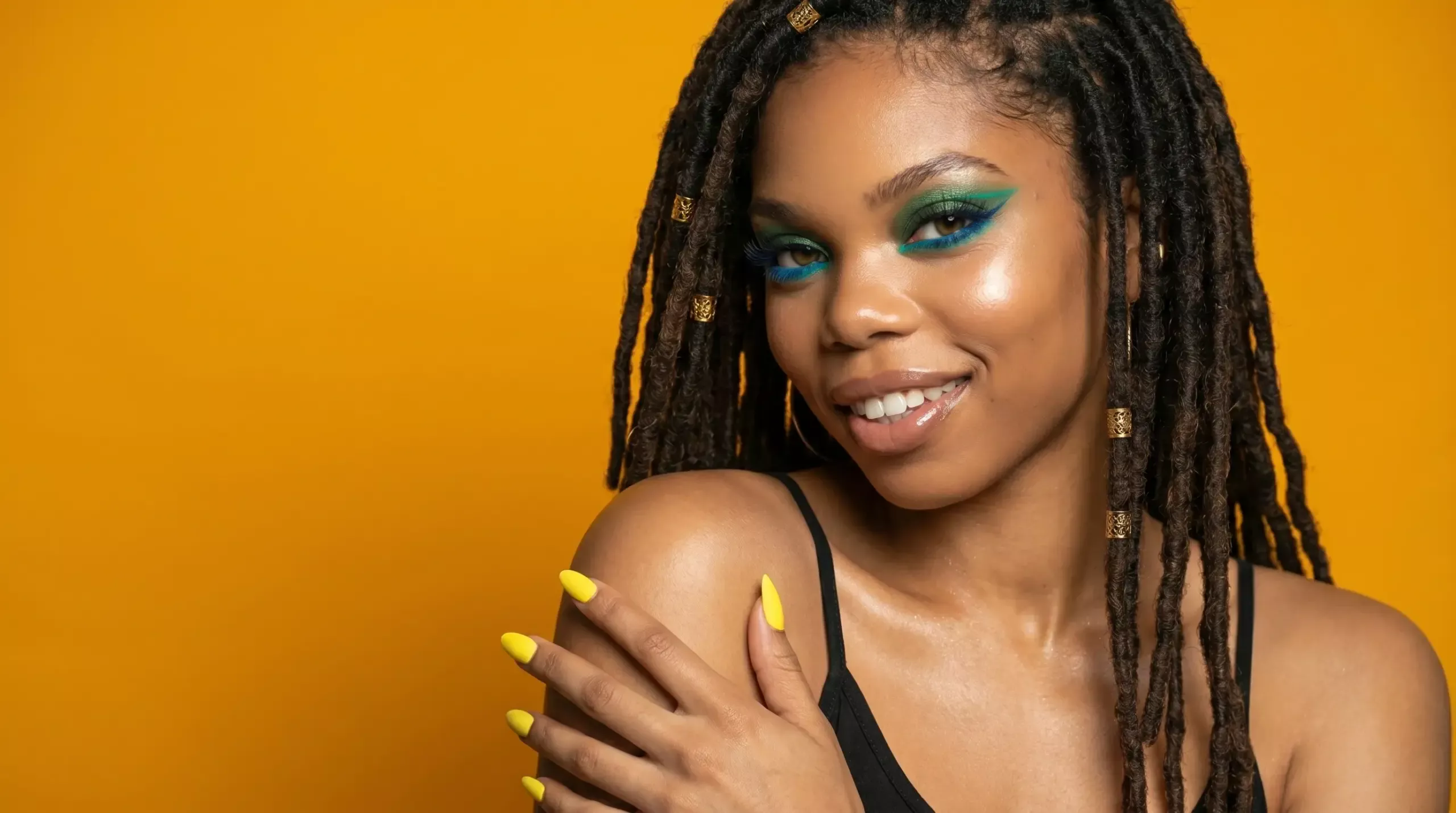 Smiling woman with long-lasting teal and green eyeliner look featuring blue lashes and yellow nails against an orange background